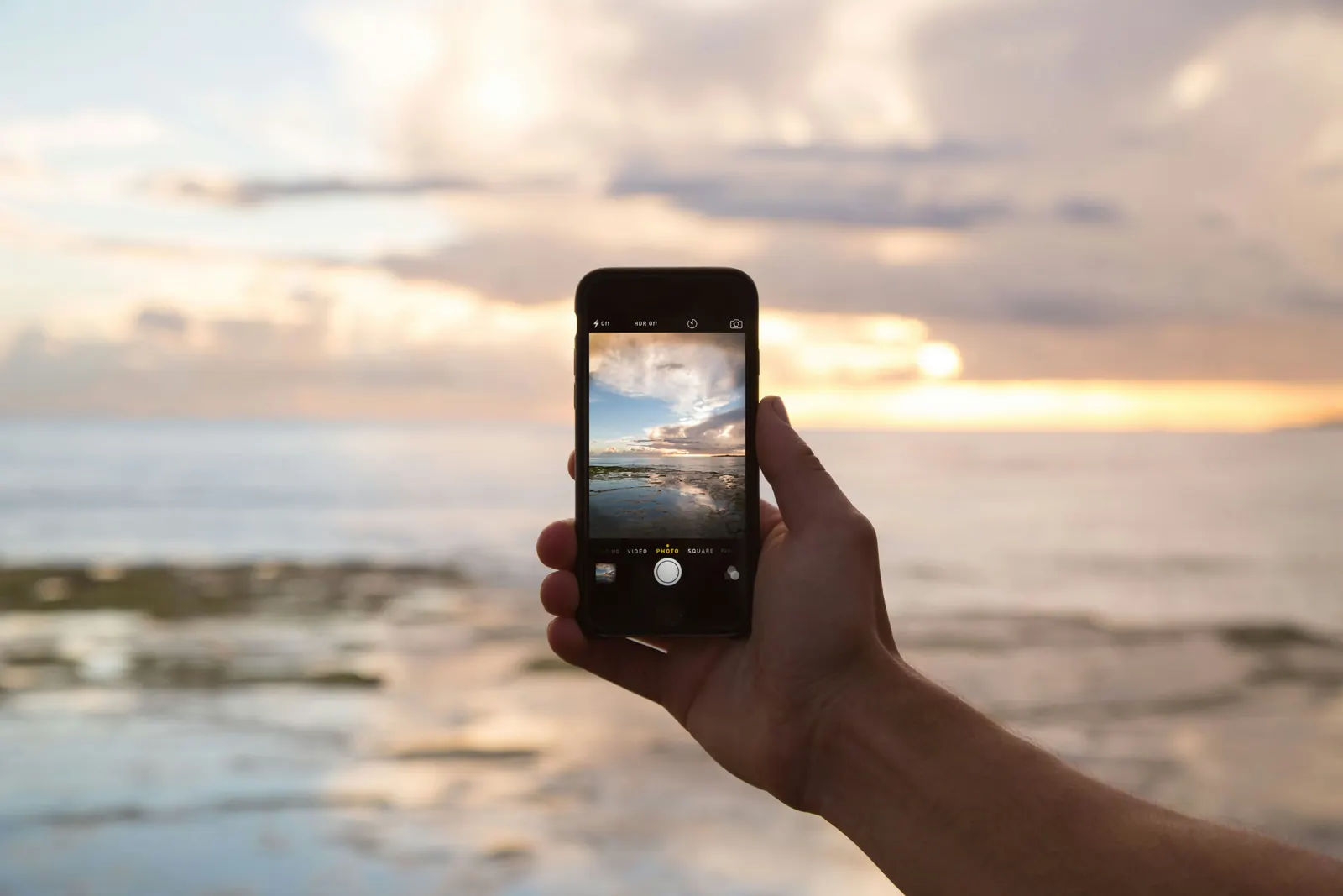 A hand holding up an iPhone to photograph a sunset over the sea.